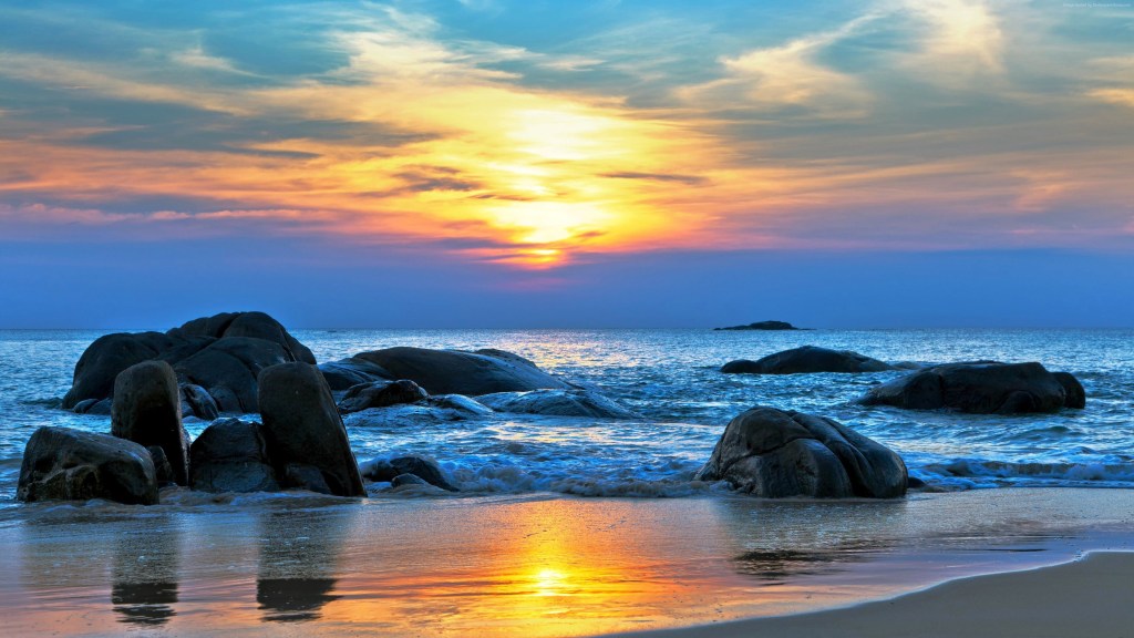  A serene beach at sunset featuring large, dark rocks scattered along the shoreline. The ocean reflects vibrant hues of orange, yellow, and blue from the colorful sky. Gentle waves lap against the rocks, creating a peaceful and tranquil atmosphere. This picturesque setting is surrounded by an open horizon, emphasizing the vastness of the sea and sky.