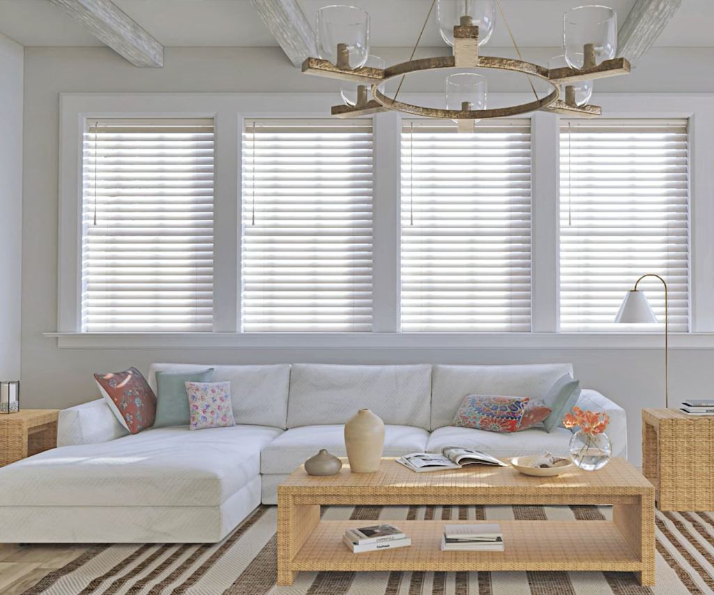 A bright and airy living room with white wooden blinds on a series of windows, diffusing natural light into the space. The room includes a white sectional sofa, colorful patterned throw pillows, and a woven coffee table, creating a casual yet elegant atmosphere.