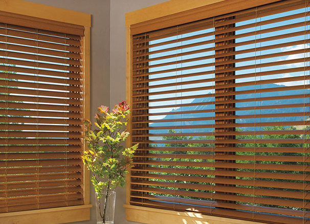 Two large windows with wooden blinds, allowing a scenic view of a lush green landscape and distant mountains. The blinds feature a warm wood tone, complementing the wooden window frames and cozy interior.