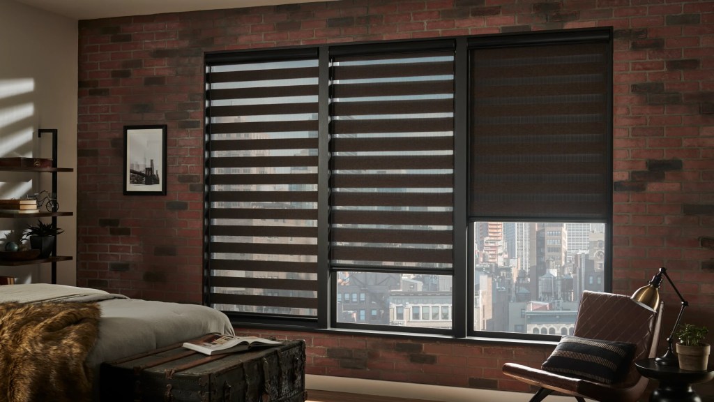 An industrial-style bedroom with exposed brick walls and large windows covered by dark zebra blinds. The blinds are partially open, revealing a cityscape view. The room is furnished with a rustic wooden trunk as a table, a leather chair, and a cozy faux fur blanket on the bed.