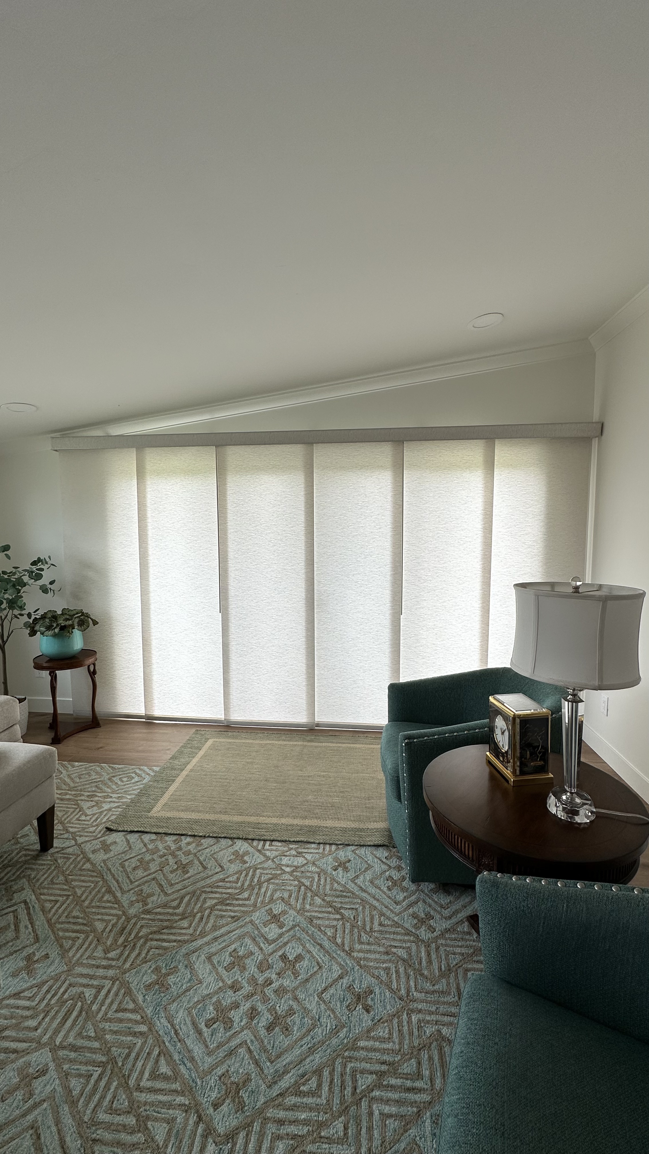 A bright and elegant living room with vertical panel shades in a soft white color covering a large sliding glass door. The room is styled with a teal armchair, a round wooden coffee table, and a side lamp, creating a cozy and sophisticated atmosphere.