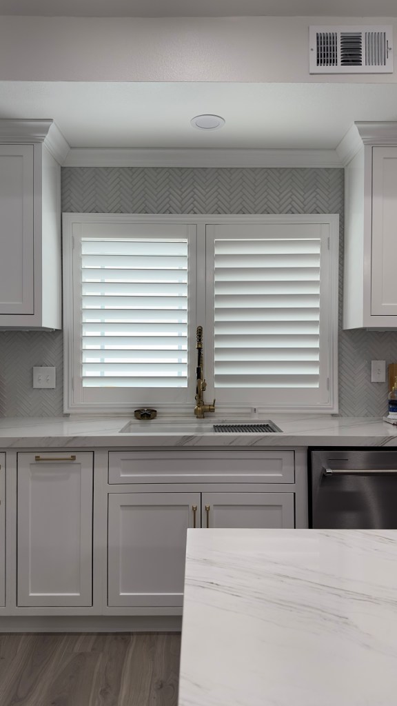 A kitchen window outfitted with white plantation shutters, adding a sleek and functional design element. The shutters sit above a stainless-steel sink and are surrounded by classic white cabinetry with a modern backsplash.
