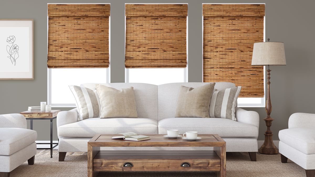 A cozy living room featuring warm brown woven wood shades on three large windows. The shades complement the beige walls and soft gray sofa. The wooden coffee table and accents of greenery add an earthy, harmonious touch to the space.